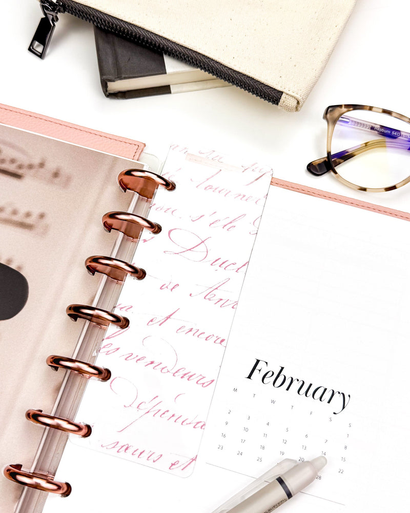 Romantic pink colored French handwriting on a glass clear plastic Page Finder. On a discbound planner with Rose Gold discs and Blush Snap-on Vegan Leather Cover on a desk flat lay. 