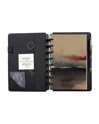 Open black pebbled leather discbound planner with interior pockets, pen loop, and black discs, featuring a removable dashboard with an abstract watercolor landscape in warm beige, charcoal, soft gray, and muted rust tones.