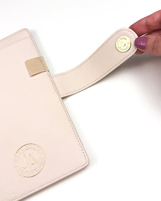 Close-up of a light beige planner cover with a textured finish, featuring a gold foil Jane’s Agenda logo and an extended snap closure strap with gold hardware. A hand holds the open strap to demonstrate the secure magnetic closure, while the coordinating elastic pen loop is visible against a clean white background.