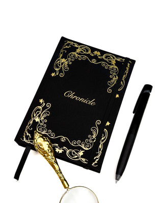 A Black And Gold Notebook With the word Chronicle On the front, with a pen and a magnifying glass to the side of it.
