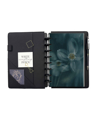 Open black pebbled planner cover with interior storage pockets, elastic pen loop, magnetic bookmark, and decorative cards. A removable floral dashboard featuring a soft blue flower on a deep teal background is displayed on black discs, with a pen secured in the built-in pen loop.