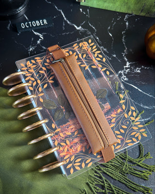 A clear planner cover with an autumn leaf pattern and a brown leather zipper pouch, shown in a styled flat-lay on a dark marble surface with green fabric and a black stamp.