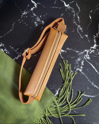 Tan zippered planner pouch with an adjustable matching strap and gold-tone hardware, displayed on a black marble surface alongside a textured green fabric with fringe.