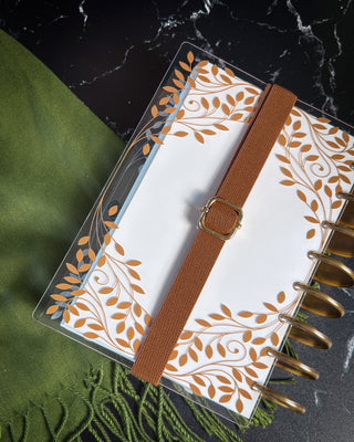 Clear discbound planner cover featuring an elegant copper leaf and vine design, secured with a brown elastic band and gold buckle. The planner has gold discs and is styled on a dark marble surface beside a textured olive green fabric.