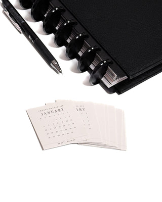 Set of twelve 2026 calendar cards with a black discbound planner and pen in the background over a white background.