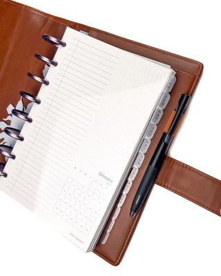 Close-up of an open discbound planner inside a warm brown leather cover, featuring a transparent frosted plastic dashboard protecting the planner pages. Frosted monthly index tabs remain visible along the right edge for easy navigation, while a black pen is secured in the leather pen loop, highlighting the dashboard’s clean, functional design.