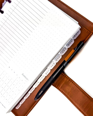 Close-up of an open discbound planner in a warm brown leather cover, showcasing transparent frosted monthly index tabs visible along the page edges. A black pen sits in the built-in pen loop, while the layered planner pages and frosted tab dividers demonstrate an organized, easy-to-navigate planner setup.