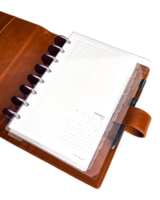 An open discbound planner in a warm brown leather cover featuring a transparent frosted plastic dashboard layered over lined planner pages. Frosted monthly index tabs run down the right side, while a pen is tucked into the built-in pen loop, showing how the clear dashboard protects pages while keeping tabs visible and accessible.