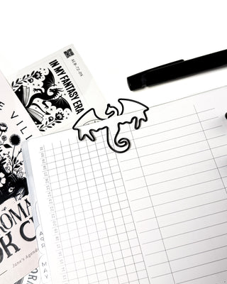 Black dragon-shaped metal paper clip clipped to a planner page, surrounded by a black pen, bookish fantasy stickers, and planner inserts. The clean workspace highlights the whimsical dragon clip as a decorative bookmark for planners, journals, or books.