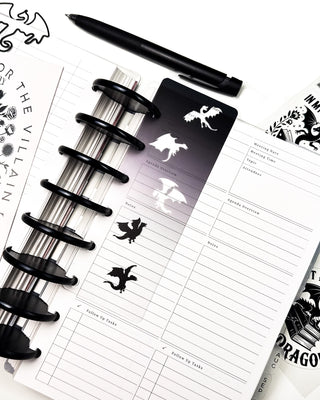 Open discbound planner featuring a transparent dragon-themed bookmark with black and white dragon silhouettes marking a meeting notes page. A black pen, dragon-shaped paper clip, bookish sticker sheet, and coordinating planner accessories surround the planner, creating a clean, fantasy-inspired workspace.
