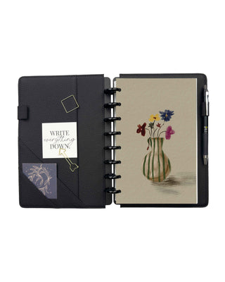 Open black pebbled leather discbound planner with interior pockets, pen loop, and black discs, featuring a removable dashboard illustrated with a green striped vase holding a colorful bouquet of hand-painted wildflowers on a soft cream background.