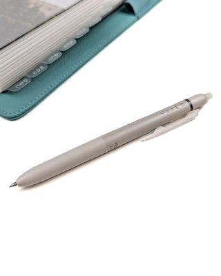 Beige retractable and erasable Pilot frixion brand pen that has black ink, a rubber grip, and .05 writing tip with a teal planner with tabbed dividers sitting in the distance on a white background