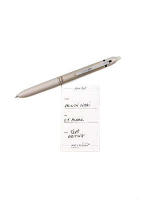 Pen writing card for ink test with a beige retractable and erasable Pilot frixion brand pen that has black ink, a rubber grip, and .05 writing tip on a white background