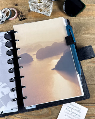 Frosted translucent planner dashboard featuring a soft golden island landscape, layered over a discbound planner to showcase its semi-transparent finish. Styled with black discs, a blue pen, black leather planner cover, sunglasses, washi tape, and desk accessories on a wooden desk.