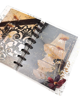 Open Jane’s Agenda discbound planner with dark metallic discs, featuring layered clear dashboards with ornate black scrollwork, gold handwritten script, glowing crystal chandeliers, delicate pressed flowers, and a deep burgundy bloom.