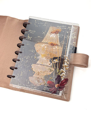 Open Jane’s Agenda discbound planner with dark metallic discs inside a bronze textured cover, featuring layered clear dashboards with glowing crystal chandeliers, gold handwritten script, delicate pressed flowers, and a deep burgundy bloom.