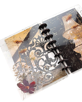 Open Jane’s Agenda discbound planner with dark metallic discs, featuring layered clear dashboards with handwritten script, delicate pressed flowers, a burgundy bloom, ornate black scrollwork, and glowing crystal chandeliers.
