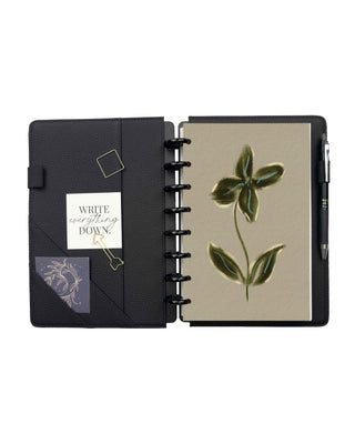 Open black planner cover with a textured finish featuring interior storage pockets, card slots, a pen loop, and black metal discs holding a botanical notebook. The left side displays decorative cards and paper clips, while the right side showcases the notebook securely held inside the cover.