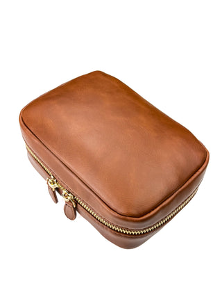Angled view of a warm brown leather mini train case displayed against a clean white background. The case features a smooth leather exterior with natural grain, rounded edges, gold-tone dual zipper hardware, and matching leather zipper pulls, showcasing its structured, compact design.
