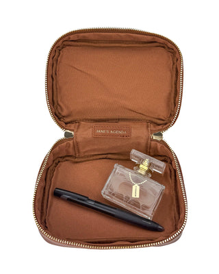 Open warm brown leather mini train case displayed on a white background, revealing a spacious brown fabric-lined interior. The case is shown holding a black pen and a small perfume bottle to demonstrate storage capacity, with gold-tone zipper hardware and a Jane’s Agenda branded interior label visible.