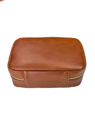 Back view of a warm brown leather mini train case displayed against a clean white background. The mini train case features a smooth leather exterior with natural grain, a convenient rear slip pocket, rounded corners, and gold-tone zipper hardware for a classic, travel-ready design.