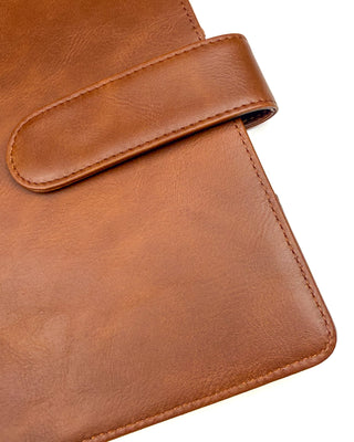 Close-up of a warm brown leather planner cover featuring the rounded snap closure strap and finely stitched edges. The detailed view highlights the smooth leather texture, rich caramel tone, and quality craftsmanship against a clean white background.