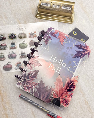 Jane’s Agenda discbound planner with dark purple discs and a clear fall leaf cover layered over a pumpkin dashboard, featuring “Hello Fall” in white lettering, styled with autumn pumpkin stickers, a pen, and a vintage desk calendar.