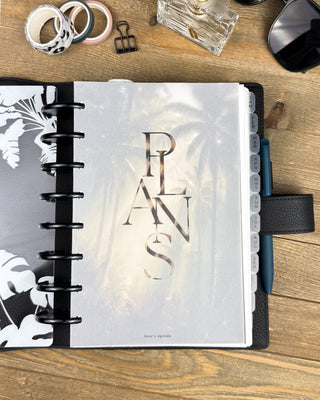 Lifestyle photo of an open discbound planner featuring a minimalist “PLANS” dashboard with modern stacked typography and a subtle tropical background. Styled on a wooden desk with a black planner cover, monthly tabs, blue pen, washi tape, binder clip, and desktop accessories, creating a clean, organized planning workspace.