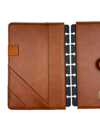Interior view of a warm brown leather planner cover shown open, highlighting two angled card pockets, a full-length slip pocket, and a coordinating elastic pen loop on the left side. The opposite side features a large document pocket with black discbound inserts visible at the center, showcasing a clean, functional layout for organizing planner pages and accessories.