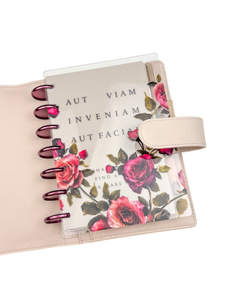 Light beige planner cover shown holding a compact discbound notebook with metallic rose-colored discs and a translucent floral cover. The planner’s interior slip pocket securely stores the notebook, demonstrating compatibility with smaller planners and accessories.