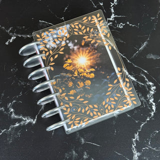 Jane’s Agenda discbound planner featuring a clear cover decorated with elegant gold botanical vines and leaves, layered over an autumn dashboard with golden sunlight shining through fall foliage. Styled with silver discs on a black marble surface with white veining.