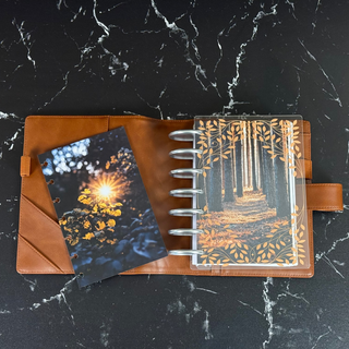 Jane’s Agenda open discbound planner featuring a clear cover decorated with elegant gold botanical vines and leaves, layered over a sunlit forest pathway dashboard. Styled in a warm brown leather cover with silver discs, alongside a coordinating autumn dashboard featuring golden sunlight shining through fall foliage, all displayed on a black marble surface.