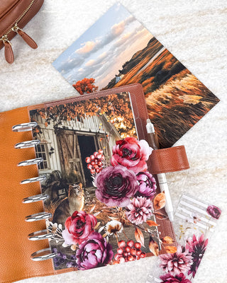 Jane’s Agenda autumn-themed discbound planner featuring a rustic cat-and-barn dashboard layered with rich burgundy, plum, and red florals, berries, and fall foliage. Styled in a warm brown leather cover with silver discs, a white pen, and a coordinating golden landscape dashboard.