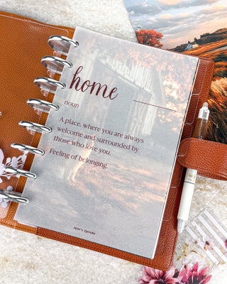 Jane’s Agenda translucent vellum dashboard featuring the word “home” and the definition, “A place, where you are always welcome and surrounded by those who love you. Feeling of belonging.” Layered over a rustic autumn cat-and-barn dashboard in a warm brown discbound planner with silver discs and a white pen.