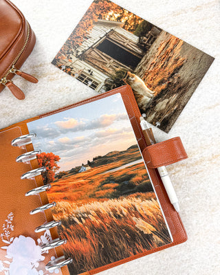 Jane’s Agenda discbound planner featuring an autumn landscape dashboard with golden grasses, orange foliage, rolling hills, and a white farmhouse beneath a soft cloudy sky. Styled in a warm brown leather cover with silver discs, a white pen, and a coordinating rustic cat-and-barn dashboard.