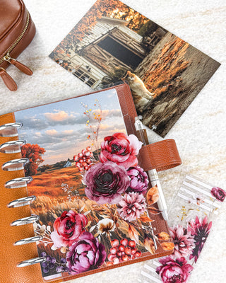 Jane’s Agenda autumn-themed discbound planner featuring a golden landscape dashboard layered with rich burgundy, plum, and red floral accents, berries, and fall foliage. Styled in a warm brown leather cover with silver discs, a white pen, coordinating floral pieces, and a rustic cat-and-barn dashboard.