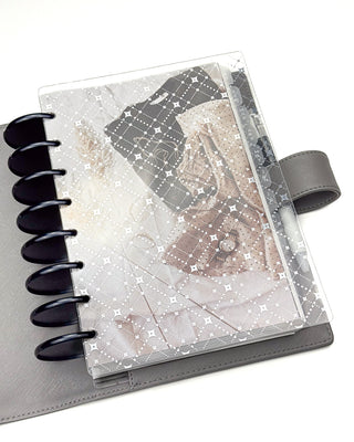 The gray leather cover with a planner featuring clothes gently laid out on white linens layered with the silver foiled diamond pattern clear cover. 