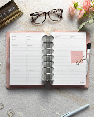 Open discbound planner with pink leather cover on a light gray granite surface, accompanied by pens, glasses, and flowers.
