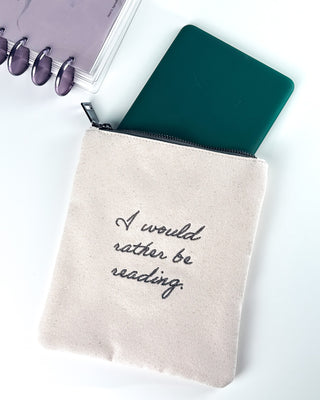 Natural canvas zippered book sleeve embroidered with “I would rather be reading,” shown with a green e-reader partially tucked inside. A lavender discbound planner with matching purple discs sits in the background on a white surface.