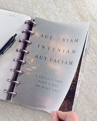 Open Jane’s Agenda discbound planner with metallic purple discs and monthly side tabs, showing a hand lifting a frosted dashboard featuring the Latin phrase “Aut viam inveniam aut faciam” and the English translation, “I shall either find a way or make one.”
