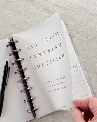 Open Jane’s Agenda discbound planner with metallic purple discs and monthly side tabs, showing a hand lifting a frosted dashboard featuring the phrase “Aut viam inveniam aut faciam” and its English translation, “I shall either find a way or make one,” with planner pages visible underneath.