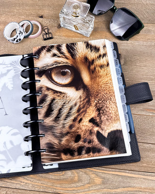 Lifestyle photo of a discbound planner featuring a striking dashboard with a close-up portrait of a leopard, highlighting its golden eye and detailed spotted fur. The planner is styled on a wooden desk with black discs, sunglasses, washi tape, a perfume bottle, and desk accessories, creating a bold, safari-inspired planning aesthetic.