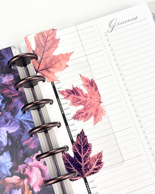 Open Jane’s Agenda discbound planner with dark metallic discs, featuring a clear page finder decorated with oversized pink and deep purple maple leaves, layered over weekly meal-planning and grocery checklist pages.