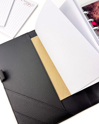 Open black textured planner cover with interior pockets, shown holding a white notebook with a kraft-colored backing, with planner accessories visible in the background.