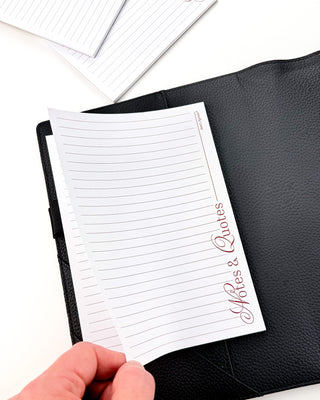 Jane’s Agenda lined “Notes & Quotes” notepad shown inside an open black textured planner cover, with a hand lifting the top sheet to reveal additional lined pages underneath.