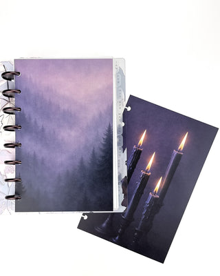 Open Jane’s Agenda discbound planner with dark metallic discs and monthly side tabs, featuring a misty purple evergreen forest dashboard alongside a coordinating dashboard with four glowing candles against a deep purple background.