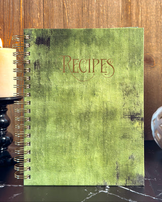 A spiral-bound recipe notebook with a distressed olive green cover and the word 'Recipes' embossed on the front, styled on a dark marble surface with lit candles and a wooden background.