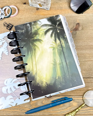 Lifestyle photo of a discbound planner featuring a lush tropical palm forest dashboard with soft rays of sunlight filtering through the trees. Styled on a wooden desk with black discs, a blue pen, magnifying glass, sunglasses, washi tape, and desk accessories for a serene, nature-inspired planning aesthetic.