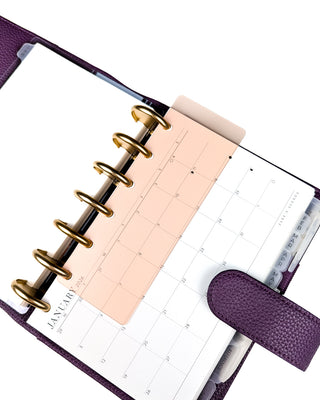 Purple discbound planner with gold binding discs and a vertical monthly calendar page with and a peach colored translucent page finder book mark on a white background