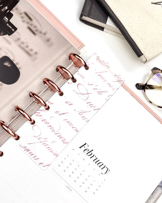 Romantic pink colored French handwriting on a glass clear plastic Page Finder. On a discbound planner with Rose Gold discs and Blush Snap-on Vegan Leather Cover on a desk flat lay. 